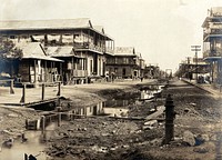 Colón, Panama, before the sanitary works that were implemented during the construction of the Panama Canal: a ditch runs down the centre of a street lined with wooden houses. Photograph, 1908.