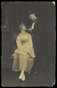 Two actors pose on stage: a man wearing top hat and tails gazes down at a man in drag. Photographic postcard, 192-.