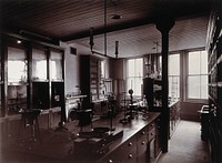The interior of a pharmaceutical or chemical laboratory, with two long benches in the centre, glass-fronted cabinets and a coal-burner along the left wall. Photograph.