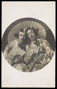 Two young men in drag wearing Japanese costumes, pose together holding fans, underneath a parasol. Photographic postcard, 1905.