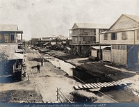 Colón, Panama: a ditch runs down the middle of the unpaved street which is lined with wooden houses; a man on horseback is in the foreground. Photograph, 1907.
