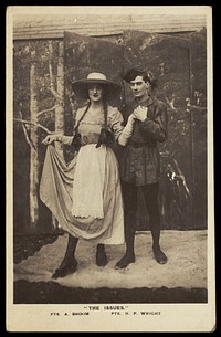 Two members of a military concert party pose on stage: one is in drag with a large hat and pigtails. Photographic postcard, 1915-1916.