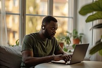 Black man typing on his laptop computer sitting window.