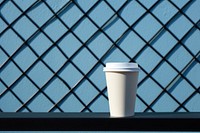 A paper coffee cup is on a black grid fence white blue wall.