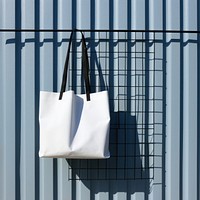 A white fabric tote bag is hanging on a black grid fence handbag wall blue.