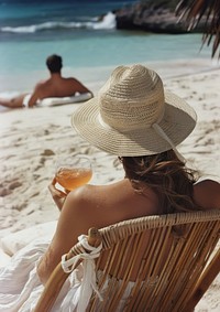 A woman sitting in a sun chair sipping a cocktail beach hat sunbathing.