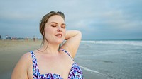 Curvy woman enjoying the beach, eyes closed, feeling the breeze. Relaxed woman at the beach, wearing a patterned swimsuit, standing by the ocean, serene expression. Curvy blonde woman at beach.