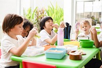 Children enjoying lunch at a green table, eating sandwiches and snacks. Kids in a bright room, sharing a meal, smiling and having fun together. Diverse young students eating school lunch.