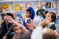 Children in a classroom raising hands. Diverse group of kids, including boys and girls, engaged in learning. Classroom setting with books and eager students. Little kids raising hands in classroom.