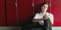 Teen student sitting by red lockers, wearing casual clothes. Student boy sitting depressed on school corridor. School boy sitting by the school locker thinking. School bullying. School boy thinking