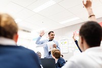 Man teacher engages students in a classroom, holding papers and raising a hand. Diverse students listen attentively, interactive learning. Man teacher in class hands up with students participate