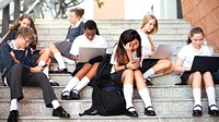 Students in uniforms sit on steps, using laptops and phones. Diverse group of boys and girls engaged in technology, studying and socializing outdoors. Students working on laptop in school.