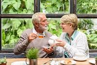 Elderly couple enjoying tea in a cozy setting. The couple, a white man and woman, share smiles and tea. Relaxed atmosphere with greenery and tea cups. Senior couple at afternoon tea date.