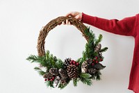 Hand holding a festive wreath with pinecones and greenery. Wreath features pinecones, greenery, and berries. Holiday wreath with pinecones and festive decor. Woman holding a Christmas wreath.