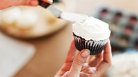 Close-up of hands frosting a cupcake with white icing. Cupcake decorating with icing. Sweet treat with icing and cupcake focus. Baking and icing in action. Vanilla frosting on chocolate cupcake.