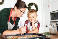 A woman and child decorate cookies in a kitchen. The woman wears glasses and an apron. The child watches eagerly. Holiday wreaths hang in the background. Mother and son baking for Christmas.