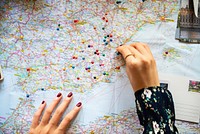 A close-up of a woman's hand, possibly placing colorful pins on a detailed map. The map features various locations, emphasizing travel and exploration.