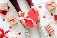 Hands exchanging a heart-shaped gift box with ribbon. Surrounded by wrapped presents and red hearts. Gift exchange, heart, and presents theme. Valentines gift of love.