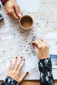 Hands on a map with colorful pins, planning a journey. Map and pins show travel ideas. Coffee cup nearby. Map, pins, and travel planning scene. Women planning a trip on a map.