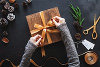 Hands tying a gold ribbon on a gift box. Gift wrapping with ribbon, pine cones, and scissors. Festive and creative gift wrapping with ribbon and decorations. Woman wrapping a festive Christmas gift.