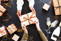 Hands exchanging a gift box with ribbon, surrounded by more gift boxes, champagne glasses, and festive decorations on a black surface. Festive holiday gifts.