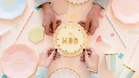 Top view of a birthday celebration setup with hands holding a cake. Pastel plates, cups, and straws surround the cake. Festive and colorful pastel birthday props in hands. Hands holding birthday cake.