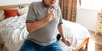 Elderly man sitting on bed, hand on chest, appearing unwell. Casual attire, bedroom setting. Concerned expression, elderly man, health issue, bedroom scene. Elderly man with chest pain, heart attack.