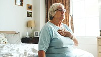 Elderly woman sitting on bed, hand on chest, in a bright room. Senior woman in a peaceful setting, wearing a light blue shirt, with framed photos in the background. Elderly woman touching chest.