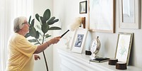 senior woman dusting a shelf with framed photos and a clock. Bright room with a plant. Dusting, cleaning, and organizing in a cozy home environment. Senior woman dusting in home.