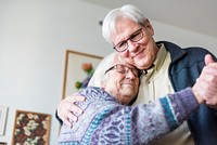 Elderly couple dancing and embracing in a cozy room. Both wear glasses and have white hair. The couple shares a joyful moment, smiling and holding each other. Happy elderly couple dancing in the room.