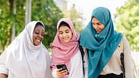 Three muslim women friends in hijabs walk together, smile and look at phone. Diverse muslim women friends group enjoy sunny day, use phone for social media, happy and cheerful Muslim women friends