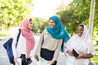 Three women walking outdoors, wearing colorful hijabs. They are smiling and holding books. Diverse group enjoying a sunny day, sharing a joyful moment. Young muslim girls walking in the park.