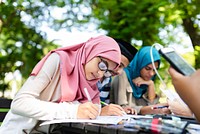 Group of students studying outdoors. Focused young woman in hijab writing. Diverse group engaged in learning. Education and teamwork in a natural setting. Female muslim teenage students and friends.