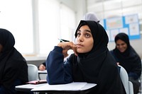 A student in a classroom, wearing a hijab, looks thoughtful. Classroom setting with students. Focused student, learning environment, education scene. Young Muslim girl wearing hijab in school.