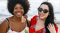 Diverse two women enjoying a beach day. African American woman wears a floral dress, the other a red floral dress. Both are smiling, with the ocean in the background. Women friends smiling at beach