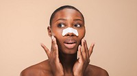 Black woman with short hair applies a nose strip, touching face. Skincare routine with a nose strip for clear skin. Neutral background enhances focus on skincare. Black woman doing skincare routine.