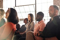 Diverse group of people sitting in a sunlit room, attentively listening. Mixed genders and ethnicities, focused and engaged in a bright, airy space. Group of people sitting together, community.