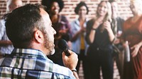 Man in wheelchair speaking to microphone in public with microphone, surrounded by diverse crowd. Joyful and supportive atmosphere, engaging speaker. Inclusive, diverse audience, lively event.