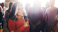 Latina woman with glasses smiles while holding a microphone in a sunlit room, surrounded by people. She wears a red top and has long, highlighted hair. Latina woman presenting in microphone at event.