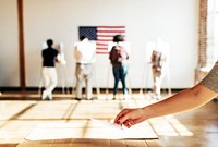 Hand placing a ballot with diverse people voting in a background with an American flag. Voting booths lined up. Hand placing a ballot. Voting scene. US election day. Vote for American democracy.