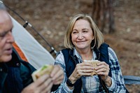 Elderly woman enjoying a sandwich while camping. Outdoors, nature, and camping vibes. Smiling and relaxed in a natural setting with a tent nearby. Mature caouple camping with tent.