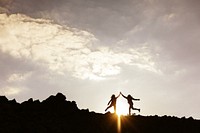 Silhouettes of two people jumping on a rocky hill at sunset. Joyful silhouettes against a dramatic sky. Energetic jump, rocky hill, and sunset glow. Hiking adventure on mountain trail.