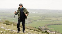 Elderly man hiking with poles on grass hill. Outdoor activity in nature. Senior enjoying hiking in nature landscape. Scenic landscape. Senior man hiking, outdoor activity in nature grass hill