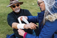 A person bandaging another's arm outdoors. The scene shows care and assistance. Bandaging and helping, supportive interaction. Senior man with injured arms while on hike in nature.