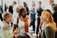 A diverse group of people, including women and men of various ethnicities, engage in lively conversation at a social event. The atmosphere is vibrant, with laughter and networking among attendees.