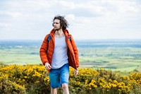 Man in orange jacket and blue shorts walking through a field. Outdoor adventure with a man enjoying nature. Bright orange jacket stands out in the landscape. Man hiking in British nature.