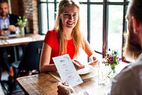 A woman in a red shirt smiles at a man in a cozy restaurant. Bright atmosphere with flowers on the table and large windows in the background. Couple dating in a restaurant.