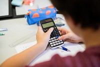 A person using a calculator at a desk with a pen and papers. The calculator is a key focus, with a blue pencil case nearby. Calculating and studying scene. Student using calculator in class.