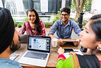 Group of young adults collaborating outdoors, using laptops and smartphones. Diverse team engaged in creative work, sharing ideas and technology. Indian startup people working together on project.