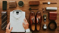 Neatly arranged men's accessories: white shirt, brown shoes, tie, wallet, watch, and grooming items on a wooden surface. Stylish men's essentials. Men's fashion and accessories top view.